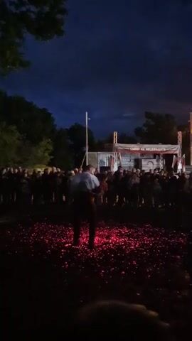 The video shows a fire-walking ceremony at night. The main subject is a man walking barefoot across a bed of glowing red embers, in front of a crowd of onlookers. The crowd is silhouetted against the dark sky, which has a deep blue hue. Behind the crowd, there's a stage with a banner written in a foreign language and what appear to be two Bulgarian flags on either side. The scene is surrounded by trees and there is a lit pole to the left. The atmosphere is solemn.