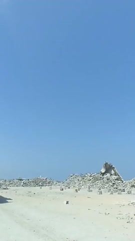 A white UNDP truck with a black grille and cage is driving on a dirt road. The truck has the number 18 in a circle on its side. There are piles of debris in the background, and the sky is clear and blue. The truck is kicking up dust as it drives.