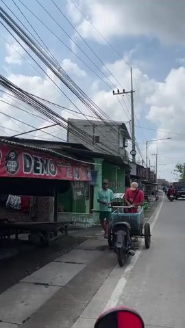 The video shows a street scene with two men on the side of the road. In the foreground, a man wearing a red shirt and a baseball cap is standing next to a three-wheeled cart attached to a motorcycle. The cart contains several items, including what appears to be trash bags. Behind him, a man in a light blue shirt is standing near a small food stall or shop. The shop has a green facade and a red banner with Indonesian text that reads "MINUMAN DINGIN KOPI ES TEM" and "SAFARI." There is a sign with "Rp16.000" indicating a price. In the background, there are several buildings, power lines crisscrossing overhead, and a street with other buildings visible. The sky is cloudy.