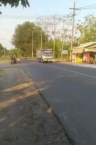 The video shows a truck driving on a paved road. Several motorcycles are also present on the road. The scene is outdoors during the daytime, with trees and power lines visible in the background. There is a small building with a sign on the right side of the road. The camera appears to be moving, capturing a view from the side of the road.