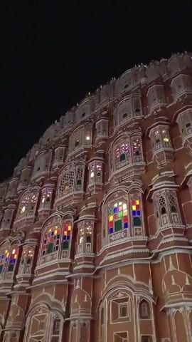 The video shows a nighttime view of the Hawa Mahal in Jaipur, India, followed by a shot of the street outside the palace. The Hawa Mahal is brightly lit, with colorful stained glass windows. The street is crowded with people, motorcycles, and cars. Some people are walking along the sidewalk, while others are taking pictures. One man is wearing a pink turban and taking a picture with his phone.
