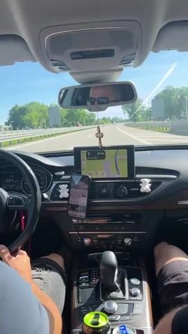 The video shows the interior of a car from the perspective of the passenger. The driver's hands are on the steering wheel, and the passenger's legs are visible in the lower right corner. The car is in motion on a highway, as indicated by the speed displayed on the dashboard (141 km/h) and the scenery passing by outside the windows. A GPS navigation system is active on the central display. There are two drink cans in the center console, one green and one blue. The overall atmosphere appears to be a sunny day during a road trip.