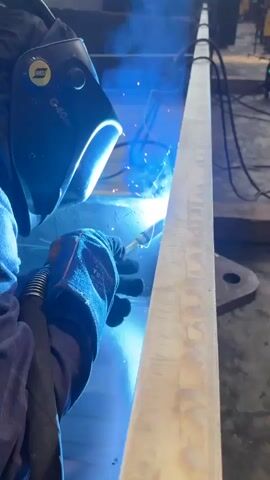 A welder wearing a blue welding mask and gloves is welding a long, thin metal beam. Sparks are flying from the welding torch and illuminating the scene with a bright blue light. The background is dark, with some indistinct shapes that appear to be machinery or other construction materials. The welder's gloved hand is holding the welding torch, and the beam is positioned vertically.