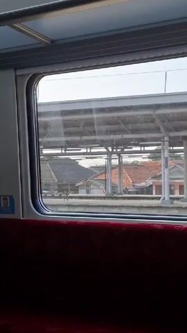 This video shows a view from inside a train as it passes through a station in Cikarang, Indonesia. The focus is on the train window, which looks out onto a train platform.

The subjects visible are primarily architectural elements of the train station. These include a covered platform with supporting pillars, roofing, and some buildings with orange tiled roofs in the background. There are also benches on the platform and what appear to be railway signals. A person is briefly visible walking on the platform. The train itself is partially visible with its red upholstered seats in the foreground. A sticker with text and a logo is visible on the train's interior wall to the left.

The activity taking place is the train moving past a stationary train station. It appears to be daytime, with bright, overcast conditions. The location is clearly identified as Cikarang, Indonesia, through external context. There are no strong emotional cues conveyed by the visual content. The overall impression is a typical view from a train window in an urban or suburban area.