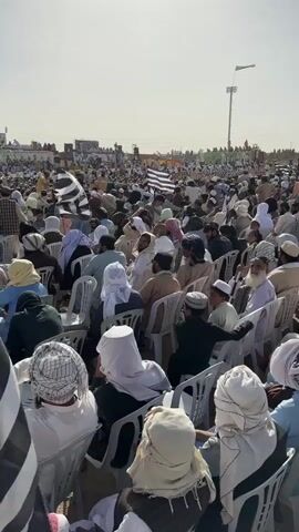 This video captures a large outdoor gathering, likely a political rally or religious event, in Pishin, Pakistan, during the daytime under a clear, bright sky. A vast crowd of men, predominantly adults but including some children, are seated on white plastic chairs, facing a stage that is partially visible on the right side of the frame. Many attendees wear traditional attire such as shalwar kameez, turbans, and prayer caps, with some also wearing white head coverings.

Numerous black and white striped flags, indicative of the Jamiat Ulema-e-Islam-F (JUI-F) party, are visible throughout the crowd and around the perimeter of the gathering. One prominent black and white flag with wide stripes is held by an individual in the foreground on the left. In the background, near the stage, a white flag with black Arabic script, resembling a Taliban flag, is also present.

The stage features large speakers on stands. Beyond the immediate seating area, more people are gathered, and in the distance, power lines or pylons can be seen against the horizon. The ground appears to be dry and dusty. The overall atmosphere suggests an organized public address or ceremony, with the crowd attentively facing the stage. A man in the bottom right corner holds a plastic bottle of water. The strong sunlight indicates mid-morning or afternoon.