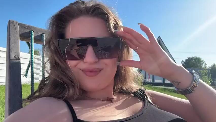 A woman with long curly hair is sitting on a wooden chair outside on a sunny day. She is wearing sunglasses, a black tank top, and a watch. There is a wooden structure and a green lawn behind her. She adjusts her hair, smiles, and poses for the camera.