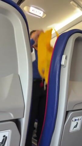 This video captures a flight attendant on an airplane, appearing to be assisting a passenger. The scene is set within the narrow aisle of an aircraft, with rows of seats on either side. The flight attendant is wearing a blue uniform shirt with a pink trim and black trousers, and has a lanyard with a badge around his neck. He is seen reaching down and pulling out a bright yellow object, possibly a blanket or safety equipment, from underneath a seat. The lighting suggests it is daytime, and the environment is typical of an airplane cabin. The flight attendant's actions seem to indicate a service or safety-related task. Visible text includes an "EXIT" sign illuminated in red above, and safety instruction pictograms on the seats, one depicting a person sitting with a seatbelt. The location context of Donja Cijevna, Montenegro is provided separately and is not visually discernible from the video itself.