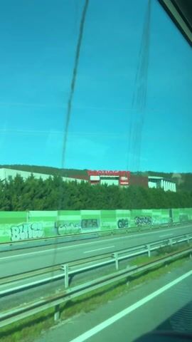 The video shows a view from inside a moving vehicle, likely a bus, looking out the window at a highway. The sky is bright blue. Across the highway is a large building with a red sign that reads "Continente." The building is partially obscured by green trees and a green noise barrier covered in graffiti. Cars and vans are seen moving on the highway. The window has some streaks on it.