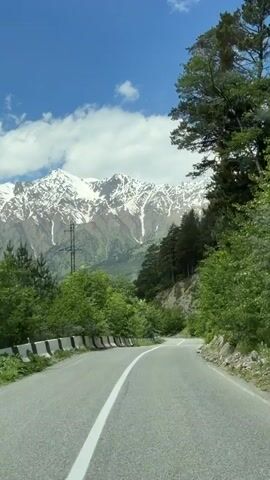 The video shows a view from a vehicle traveling on a paved road through a mountainous landscape. The road curves to the left and is lined with concrete barriers on the left side. Dense green trees and foliage flank the road, with a steep, rocky slope visible on the right. In the background, snow-capped mountains rise against a bright blue sky with scattered white clouds. Tall evergreen trees are visible on the mountain slopes. A single power line pole is visible on the left side of the road. A triangular road sign is also visible on the right side of the road. The lighting suggests it is daytime with clear weather.
