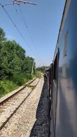 The video shows a train moving along a railway track. The perspective is from inside the train, looking out the side window. The train is moving through a rural or semi-rural area with trees and vegetation on either side of the tracks. Overhead power lines are visible. The sky is clear and blue. The train cars are dark blue and appear to be made of metal. The tracks are made of gravel and metal rails. The overall impression is one of travel through a natural landscape.