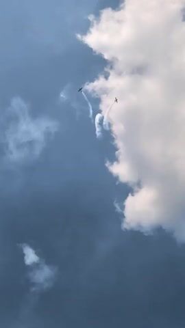 Two biplanes are performing aerobatics in a cloudy blue sky. One plane, on the left, is leaving a white smoke trail as it dives downwards and to the right. The other plane, on the right, is further up and to the right, also leaving a white smoke trail as it appears to be climbing or banking. Large white clouds are prominent on the right side of the frame, partially obscuring the blue sky.