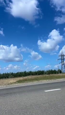 The video shows a view from a moving vehicle looking out a window at a rural landscape. In the foreground, a grey asphalt road with a white dashed line is visible. Beyond the road, there is a dry, grassy field with some bushes and trees. In the background, a tall, dark metal power line tower stands against a bright blue sky dotted with white, fluffy clouds. The camera appears to be panning slightly or the vehicle is moving, causing the landscape to shift. The lighting suggests it is daytime, likely sunny.