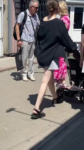 The video shows a sunny outdoor market scene. A man in a plaid shirt and sunglasses walks on the left. To his right, a woman in a bright pink dress walks away from the camera. In the center, two young women walk away from the camera, one in a black oversized shirt and white shorts, the other in a black t-shirt and white skirt. The woman on the left is pushing a stroller. The ground is paved with bricks. In the background, there are market stalls and other people walking. The overall atmosphere is casual and sunny.