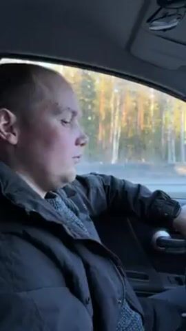 The video shows a man driving a car. He's wearing a dark jacket over a patterned sweater. The camera is positioned inside the car, focusing on the man from the passenger seat. Outside the window, trees blur by, suggesting the car is moving at a moderate speed. The lighting indicates it's daytime. The man is looking ahead, seemingly focused on driving. The interior of the car appears clean and functional.