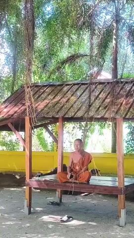 A man in an orange robe is sitting cross-legged on a wooden platform under a thatched roof. He is holding a small, dark object in his hands and appears to be looking at it. The platform is supported by wooden posts and is located outdoors in a sandy area. In the background, there is a yellow wall and lush green trees. Hanging vines are visible in the foreground. The man is the sole human figure in the scene.