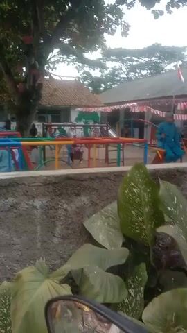 The video shows an outdoor playground with children playing on colorful climbing equipment. A brick building with a tiled roof is visible in the background, along with trees and foliage. There are several children and adults present, some interacting with the playground equipment. The overall impression is of a bright, active play area.