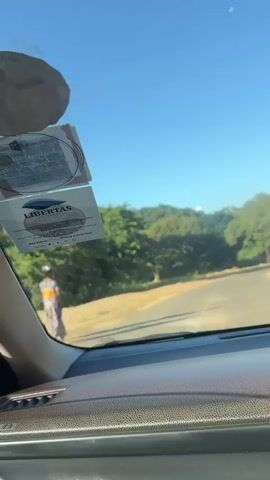 The video shows a car driving along a narrow, paved road. The sky is bright blue and clear, and the surroundings are green with many trees. The car's dashboard and a sticker on the windshield are visible in the foreground. Sand is visible on either side of the road.