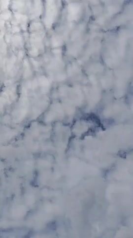 This video shows a low-angle view of a cloudy sky. The clouds are white and gray, with some darker patches indicating shadows. The clouds appear to be thin and scattered, suggesting a partly cloudy day. There is no visible horizon or ground, indicating the camera is pointed directly upwards. The lighting suggests it is daytime, but the density of the clouds makes it impossible to determine the exact time of day or weather conditions beyond cloud cover.