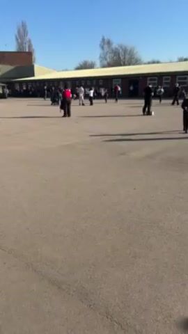 The video shows a school playground on a sunny day. A large, single-story building with many windows forms the backdrop. The playground is mostly empty but scattered with people, including children and adults. Some individuals are walking, while others appear to be playing with a ball. Shadows from the people and the building stretch across the paved surface. The sky is clear and blue, with a few trees visible in the distance.