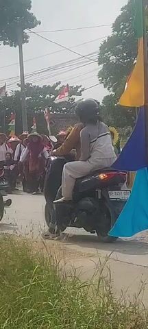 A group of people are riding motorcycles in a parade. Many of them are wearing hats or costumes. Several Indonesian flags are visible, waving in the breeze. The weather appears to be sunny and warm. The road is paved, and there are trees and buildings in the background. The video appears to be taken from a stationary position on the side of the road.