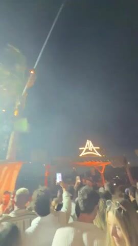 A crowded outdoor event at dusk or night features a stage with a prominent illuminated logo and lights. A palm tree with string lights is visible on the left. People are gathered in front of the stage, many holding up phones. The atmosphere appears to be that of a concert or festival.