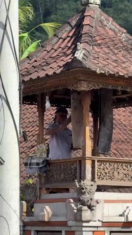A man in a light-colored shirt and a dark cap stands in a small, open-air structure with a tiled roof. He is holding a long, light-colored object, possibly a piece of wood or bamboo, and appears to be striking or cleaning it with a dark object. To his left, there is a basket or bag filled with what appear to be white flowers, possibly lotuses. The structure has intricate wooden carvings on its railings and is decorated with stone sculptures at its base, one resembling a mythical creature. Behind the structure, lush green foliage and a hillside are visible. A white electrical cord hangs down on the left side of the frame. The lighting suggests it is daytime, possibly morning or afternoon.