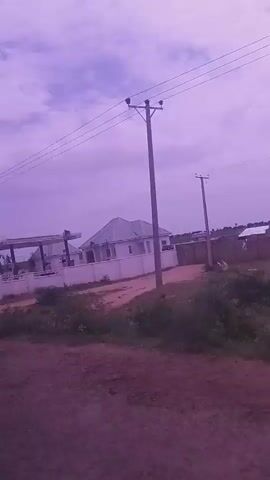 The video pans across a scene with a gas station and a white house with a fence. Power lines are visible against a cloudy, purple sky. In the distance, more power poles and sparse vegetation can be seen. The ground is reddish-brown dirt.