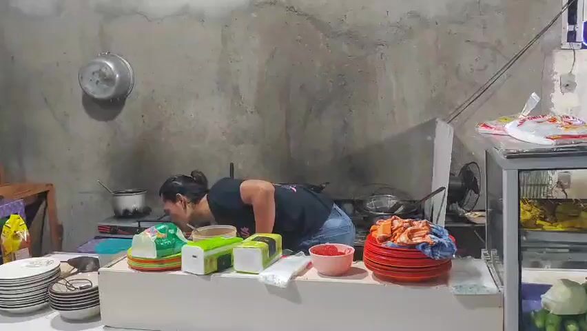 A person wearing a black t-shirt with a colorful graphic on the back and blue jeans is standing in a kitchen. The person is facing away from the camera and appears to be cooking at a stove. There is a pot on the stove, and the person is holding a utensil. In front of the person is a counter with various items on it, including plates, bowls, and napkin dispensers. To the right of the counter is a glass display case filled with food. The walls of the kitchen are concrete.