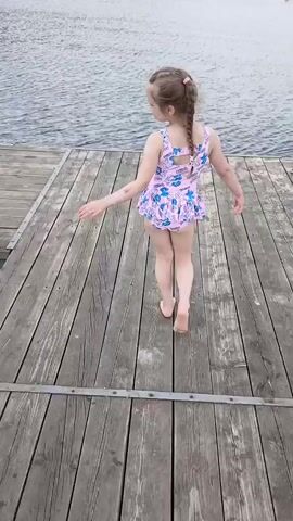A young girl in a pink swimsuit with a ruffled skirt stands on a wooden dock. She has blonde hair in a braid and is posing with her arms outstretched. She is facing away from the camera and looking to her left. The dock is made of weathered wooden planks. Behind her is a large body of water, likely a lake, with ripples on the surface. In the distance, across the water, there are trees and some buildings, suggesting a natural setting near a populated area. The sky appears overcast, indicating a possibly cool or cloudy day. The girl's expression is not clearly visible, but her posture suggests a playful or performance-like activity.