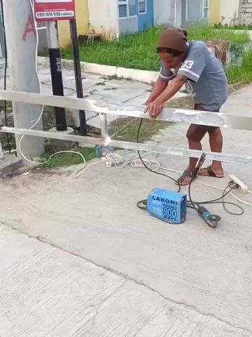 A person is welding a metal railing outdoors. The person, wearing a brown beanie and a grey t-shirt, is bent over, holding a welding torch to the railing. Sparks are flying from the welding point. A welding machine, labeled "LAKONI 900 WATT," is on the ground near the railing, along with an angle grinder and various cables. The railing is attached to a grey pole on the left side of the frame. Behind the railing, there is a patch of grass and a building. The ground in front of the railing is a concrete pathway.