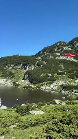 A red helicopter is flying over a mountain lake on a clear sunny day. The helicopter is a rescue helicopter. The lake is surrounded by green vegetation and rocks. The sky is blue.