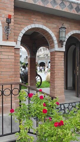 A short video clip showing a view through an arched brick entryway. Beyond the archway, a courtyard is visible with a few people interacting. A large green bush with pink roses is on the right side of the frame.