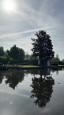 The video shows a calm body of water reflecting the sky and the scenery along its banks. A small, light-colored building sits along the water's edge, partially obscured by a large tree with dark foliage. The tree's reflection is prominent in the water. As the view pans slightly to the right, more trees and vegetation are revealed along the bank, interspersed with a few boats docked near the shoreline. The sky is pale blue with scattered clouds, and the sun appears to be shining brightly, contributing to the overall serene atmosphere of the scene.