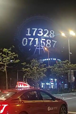 A red taxi cab is parked on the right side of the street. Behind it, a large Ferris wheel is illuminated at night. The Ferris wheel displays white numbers in two rows: "17328" on top and "071587" below. Trees and buildings with illuminated signs are visible in the background. The taxi has a "TAXI" logo on its side and a light-up sign on its roof. The scene is nighttime and urban.