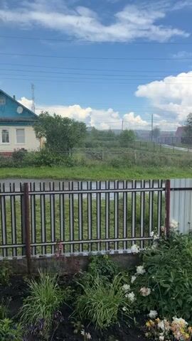 The video shows a split view of a rural landscape. The top portion of the screen features a cloudy blue sky, with power lines crisscrossing the frame. Below, a fenced-off area contains overgrown grass and bushes. In the distance, several houses and buildings are visible, along with a white car driving on a wet road. The bottom portion of the screen shows a closer view of a garden with green plants and white flowers, separated by a metal fence from the scene above. The overall impression is of a somewhat unkempt but otherwise peaceful rural setting on a day with mixed weather.