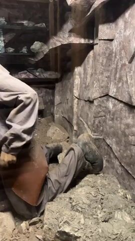 This video shows a diorama depicting miners in a rough-hewn mine shaft. In the foreground, a mannequin dressed in mining attire, including a grey jumpsuit, leather apron, and cap, is kneeling and appears to be working with a pickaxe. Another mannequin, also in mining clothes, is lying on the ground in the background, with debris around them. The environment is characterized by rough rock walls and a wooden ceiling. In the background, there is a display case filled with rocks and minerals, suggesting this is an exhibit. The video also briefly shows a wooden hallway with a display cabinet filled with items.