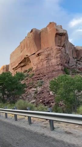 A video shows a car traveling on a road next to a large rock formation. The rock formation is a towering, reddish-orange cliff with visible layers and cracks. In front of the cliff, there are green trees and bushy vegetation. The road has a white guardrail along the side. The sky is partly cloudy with a mix of blue and gray. The video is a continuous shot from the perspective of someone inside the moving vehicle.
