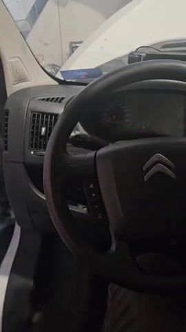 This media file captures a close-up, panning view of the interior of a vehicle, specifically focusing on the dashboard and steering wheel. The scene is dimly lit, suggesting it might be indoors or during low-light conditions. The vehicle appears to be a commercial van or truck, as indicated by the prominent gear shift lever on the center console, labeled with gear positions R, 1, 3, 5 and 2, 4, 6.

The dashboard features a black steering wheel with the Citroën logo prominently displayed in the center. Various controls are visible on the steering wheel, including buttons for what appear to be media or cruise control functions. To the left of the steering wheel, an air vent is visible, along with a speaker grille. The instrument cluster, with its gauges and needles, is also visible behind the steering wheel.

The central part of the dashboard includes a digital display screen, likely for a radio or navigation system, surrounded by buttons and knobs. Below the screen are climate control dials and other buttons. A bright yellow screwdriver is placed on the dashboard, possibly indicating that some work is being done on the vehicle. The overall impression is one of a functional, work-oriented vehicle interior. The image is static, offering no clues about the time of day or weather outside. There is no visible text other than the gear shift pattern and the Citroën logo. The location context of Bemmel, Netherlands, is provided externally.