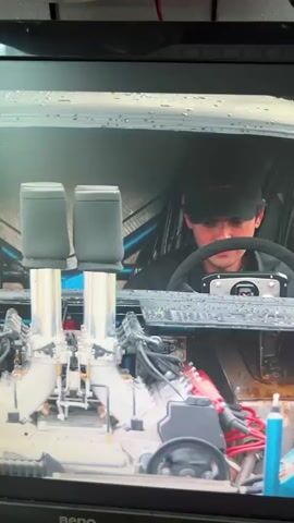 The video begins with a close-up view of a car engine with two air intakes. A person wearing a cap is visible in the driver's seat, looking down. The camera then pulls back to reveal a man in a black jacket and pants holding a small dog, standing next to the open driver's side door of the car. The car appears to be a vintage model, with a roll cage visible inside. The scene is outdoors, with green grass and trees in the background.