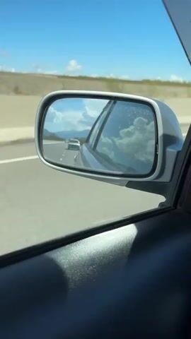The video shows a car's side mirror view while driving on a highway. In the reflection, a white car is visible on the road behind, with mountains in the distance under a blue sky with scattered white clouds. The car is traveling on a multi-lane highway. The weather appears clear and sunny. The focus is on the side mirror and what is reflected in it. The interior of the car is visible in the foreground, particularly the dark textured panel of the door.