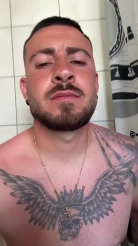 A shirtless man with a beard, short dark hair, and a tattoo on his chest stands in a bathroom with white tiled walls. He wears a gold chain necklace and black hoop earrings. He looks into the camera, and his facial expression changes from neutral to smiling, then to a full, toothy grin. A black and white patterned shower curtain is visible in the background.