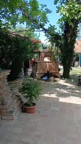 A person is walking past a brick structure with an animal figurine on top in a courtyard. The courtyard has a tiled floor and is surrounded by trees and buildings. There are tables and chairs set up on a lawn in the background.
