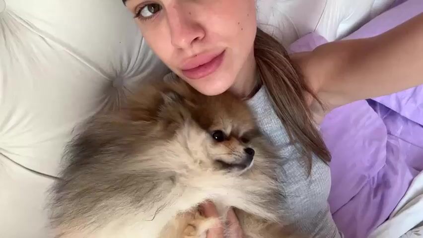 The video shows a close-up, selfie-style shot of a person lying in bed with a small, fluffy dog. The person is looking directly at the camera with their mouth open. The dog, a Pomeranian, is nestled in the crook of the person's arm and chest, facing to the right. The person is wearing a gray tank top, and the background shows a white headboard and a purple blanket. The lighting is bright, suggesting daytime.