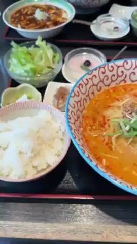 The video showcases a dining table set with a variety of dishes. The focal point is a bowl of orange-colored ramen topped with greens, placed next to a small bowl of white rice. Other items visible include a small plate with what appears to be dumplings, a bowl of green salad, a small dish of pink jelly, and a bowl of mapo tofu. There are two glasses of water in the background. The dishes are arranged on a black tray with a wooden frame. The camera pans slightly to provide a view of the various dishes.