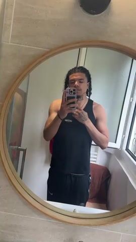 A young man takes a selfie in a bathroom mirror. He is wearing a black tank top and black Nike shorts, and has cornrows with wavy, loose ends. The man is wearing a silver chain, a black bracelet, and a ring. He raises his middle finger to the camera. The bathroom has beige tiles and a round mirror with a wooden frame. A window and a towel rack are visible in the background.