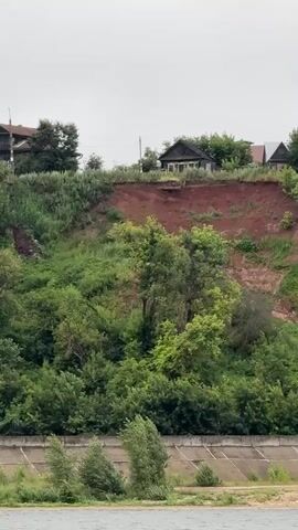 The video shows a wide river with choppy, gray water in the foreground. On the far bank, a steep, reddish-brown cliff rises up, covered in lush green trees and bushes. Atop the cliff, several rustic wooden houses are visible, nestled amongst more trees. The sky is overcast, suggesting a cloudy or possibly rainy day. The overall setting appears to be a rural or semi-rural area in Kozmodemyansk, Russia, with a natural landscape dominating the scene. There is no discernible activity taking place other than the gentle movement of the water and the stillness of the landscape. The focus is on the natural beauty of the river and the contrasting man-made structures on the cliffside. The image evokes a sense of peacefulness and perhaps a touch of isolation due to the remote setting and the overcast weather. There is no visible text in the media file.