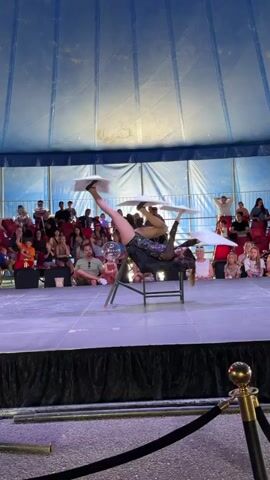 A female performer in a sparkly black costume is balanced on a chair, with her legs extended. She is balancing three white plates on her feet and hands, one on her right foot, one on her left foot, and one on her right hand. The performer is in a circus tent, with an audience watching from bleacher seats in the background. The tent has a blue and white striped fabric ceiling and white fabric walls. The stage is black and there is a black rope barrier in the foreground.