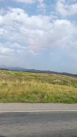 The video shows a view from a vehicle moving along a road. The road surface is visible in the foreground. Beyond the road, there is a landscape with green and yellow vegetation, including fields and some trees. In the distance, there are rolling hills or mountains. The sky is partly cloudy with white clouds against a blue sky. A utility pole with wires is visible on the left side of the frame at one point. The scene appears to be a rural or semi-rural area.