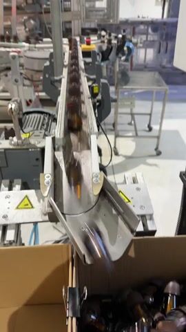 A conveyor belt moves small amber bottles toward a chute that directs them into a cardboard box. The bottles are moving quickly, causing them to appear blurred in the video. The conveyor belt is part of a larger industrial machine, with other machinery and equipment visible in the background. The environment appears to be a factory or warehouse setting.