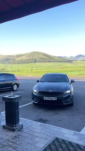 A dark-colored Kia sedan is parked in a lot with a mountainous, green landscape in the background. The car is the primary focus, with its headlights illuminated. To the left, a dark SUV is partially visible. In the foreground, a dark, stone structure with a fountain is present, along with a tiled patio and a mat. The sky is clear and blue, suggesting a pleasant day. The rolling green hills in the background are dotted with trees and fields, with a clear division between agricultural land and natural terrain. The sun appears to be low in the sky, possibly indicating early morning or late afternoon. The overall scene is serene and captures a moment of stillness in a scenic, rural setting. The license plate on the Kia is visible and reads "B 111 MY 45".